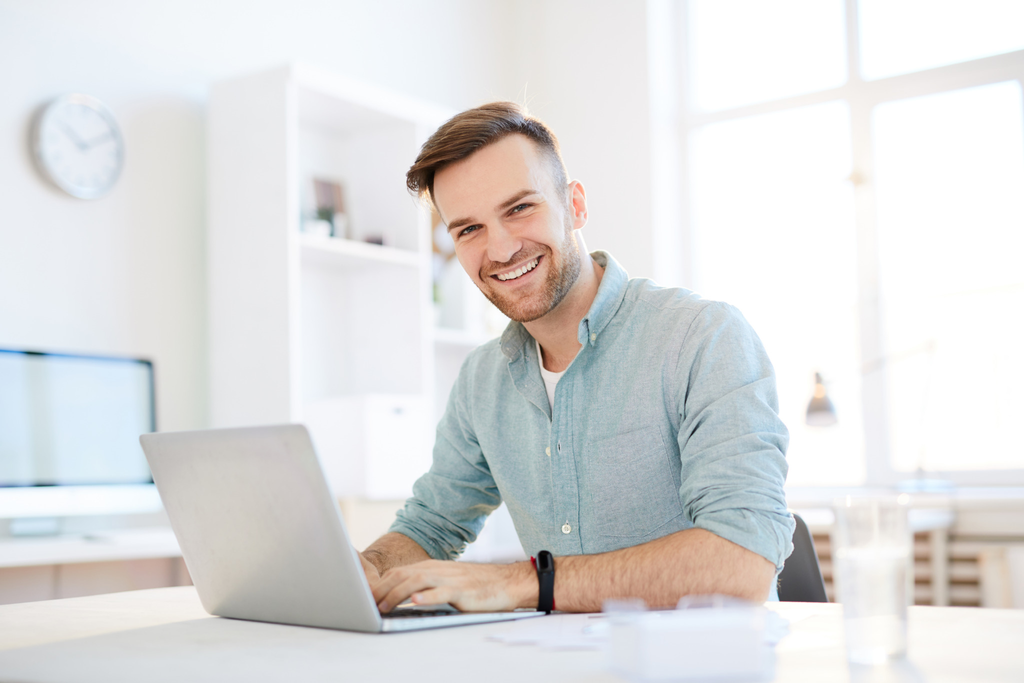 Portrait of handsome young man smiling at camera while using laptop in office, copy space