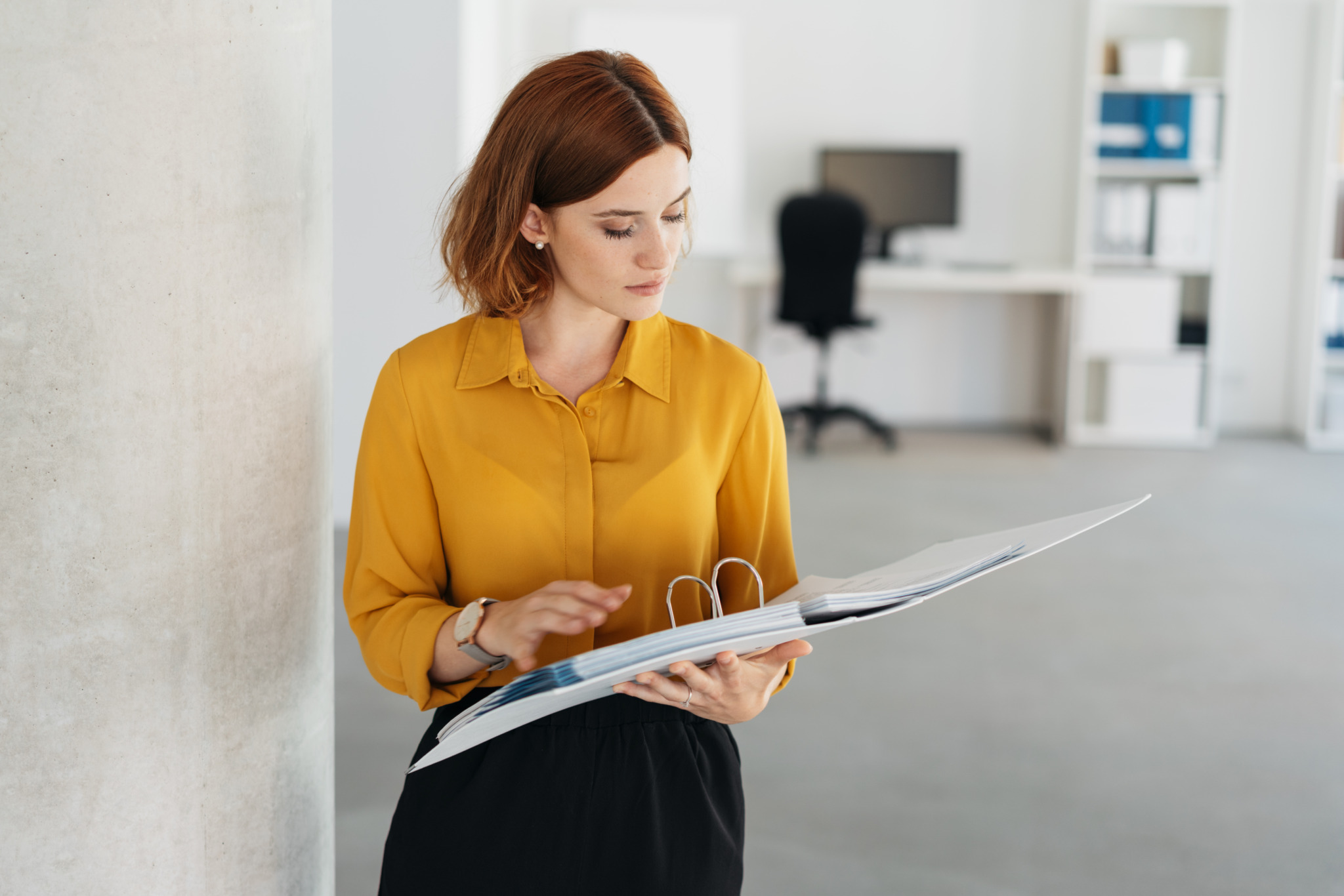 Young businesswoman consulting a large handheld file as she pauses alongside a wall in the office