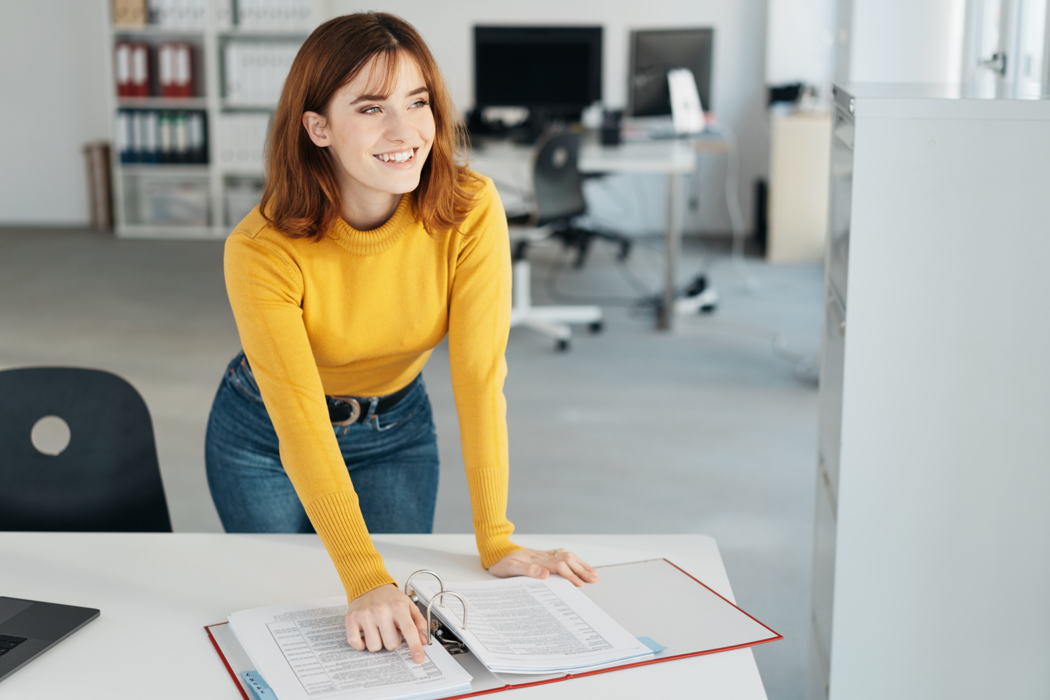 Young businesswoman interrupted in her work looking up with a smile as she marks her position on her notes with her finger
