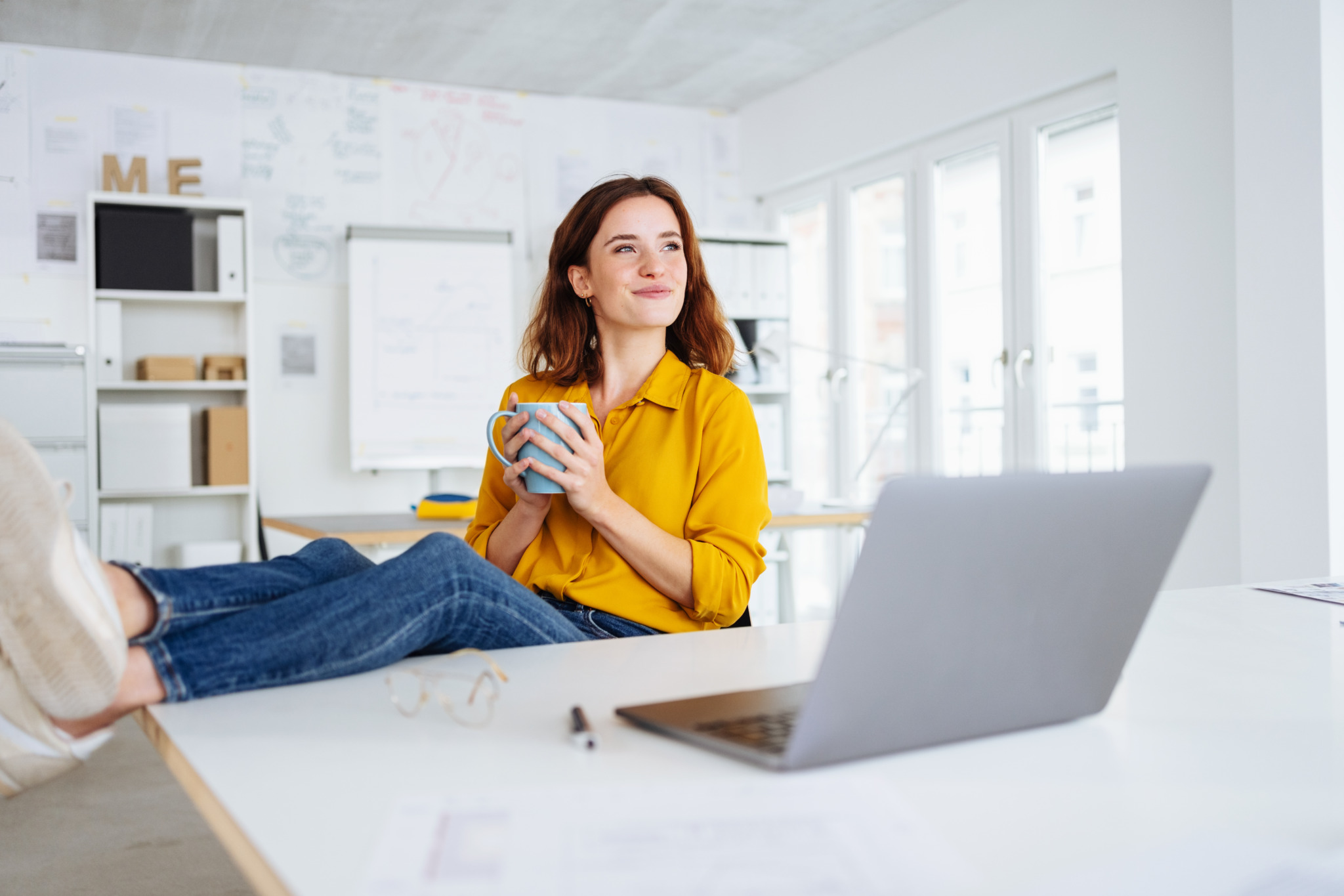 Happy smiling young businesswoman sitting daydreaming in the office as she relaxes with a mug of coffee and her feet up during a morning break