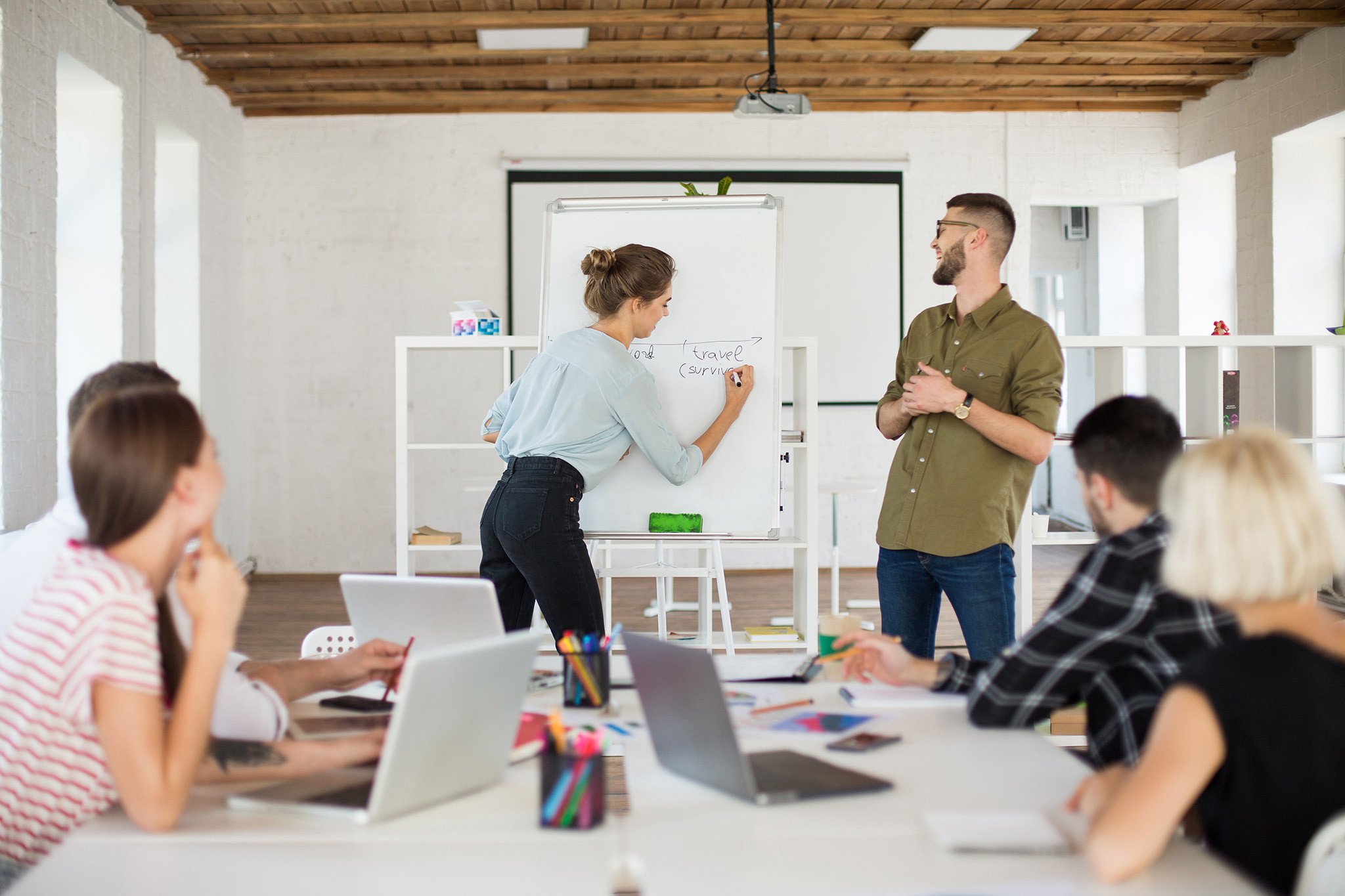 Junger Mann mit Brille und Hemd und Frau in Bluse stehen an einer Tafel und stellen Kollegen fröhlich ein neues Projekt vor Gruppe von Kreativen, die in einem modernen weißen Büro zusammenarbeiten